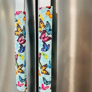 May include: Two fabric handle covers with a light blue background and a colorful butterfly pattern. The butterflies are in shades of pink, yellow, orange, blue, and green. The covers are on a stainless steel refrigerator.
