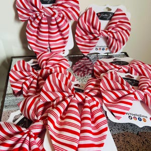 May include: Several red and white striped fabric bows are displayed on white cardstock tags. The bows are arranged in a group, showcasing their candy cane-like pattern. The tags have the text "Yoella Zima" and social media icons.