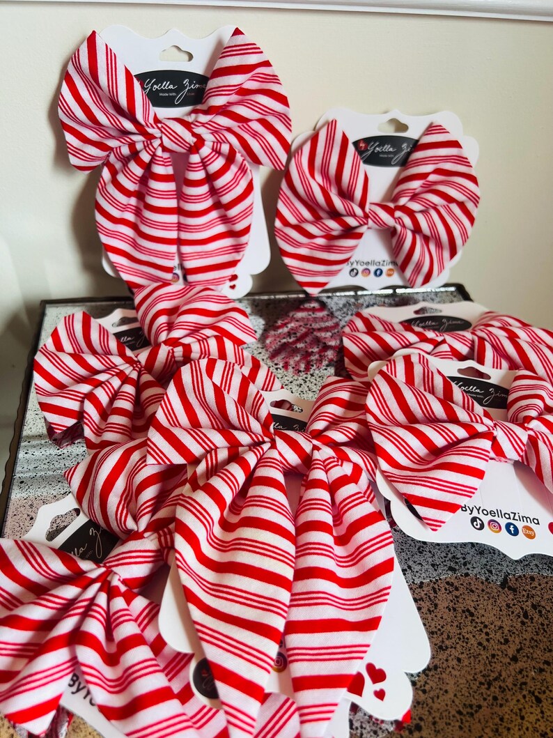 May include: Several red and white striped fabric bows are displayed on white cardstock tags. The bows are arranged in a group, showcasing their candy cane-like pattern. The tags have the text "Yoella Zima" and social media icons.