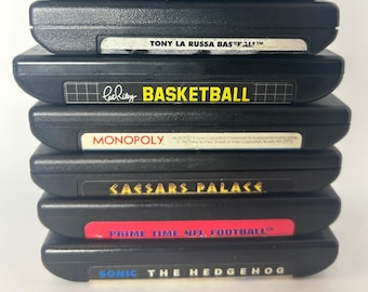 May include: A stack of eight black Sega Genesis game cartridges. The cartridges are stacked vertically, with the labels facing the viewer. The labels feature titles such as "Super Off-Road", "NHLPA Hockey 93", "Tony La Russa Baseball", "Basketball", "Monopoly", "Caesar's Palace", "Prime Time NFL Football", and "Sonic the Hedgehog".