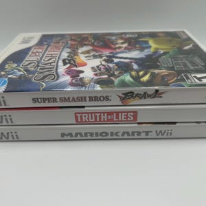 K&ouml;nnte beinhalten: Drei Wii-Spielh&uuml;llen &uuml;bereinander gestapelt. Die oberste H&uuml;lle ist "Super Smash Bros: Brawl", die mittlere H&uuml;lle ist "Truth or Lies" und die untere H&uuml;lle ist "Mario Kart Wii". Die H&uuml;llen sind wei&szlig; mit farbenfrohen Spielgrafiken.