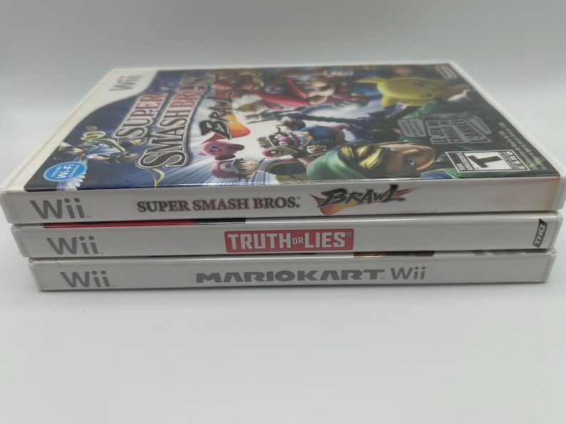 K&ouml;nnte beinhalten: Drei Wii-Spielh&uuml;llen &uuml;bereinander gestapelt. Die oberste H&uuml;lle ist "Super Smash Bros: Brawl", die mittlere H&uuml;lle ist "Truth or Lies" und die untere H&uuml;lle ist "Mario Kart Wii". Die H&uuml;llen sind wei&szlig; mit farbenfrohen Spielgrafiken.