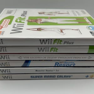 K&ouml;nnte beinhalten: Ein Stapel von Wii-Spielh&uuml;llen, darunter "Wii Fit Plus", "Wii Fit", "The Legend of Zelda: Twilight Princess", "Wii Sports Resort", "Monster Hunter Tri" und "Super Mario Galaxy". Die H&uuml;llen sind wei&szlig; mit farbenfrohen Grafiken und Text.