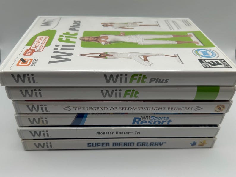 K&ouml;nnte beinhalten: Ein Stapel von Wii-Spielh&uuml;llen, darunter "Wii Fit Plus", "Wii Fit", "The Legend of Zelda: Twilight Princess", "Wii Sports Resort", "Monster Hunter Tri" und "Super Mario Galaxy". Die H&uuml;llen sind wei&szlig; mit farbenfrohen Grafiken und Text.