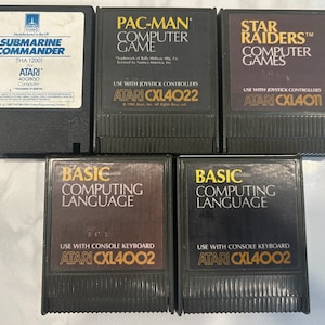 Könnte beinhalten: Fünf Atari-Spielkassetten für den Atari 400/800 Computer. Die Kassetten sind schwarz mit goldenen Schriftzügen und beinhalten Titel wie "Submarine Commander", "Pac-Man", "Star Raiders" und "Basic Computing Language".