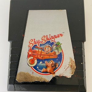 Puede incluir: Un cartucho de juego negro de Nintendo Entertainment System (NES) con una etiqueta plateada. La etiqueta presenta una ilustración de dibujos animados de un mono en un avión rojo con el texto "Sky Skipper™".