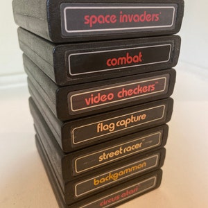 May include: A pile of seven black Atari video game cartridges. Each cartridge features a label with a game title in white or red text, including "Space Invaders," "Combat," and "Video Checkers."