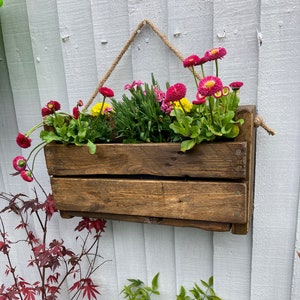 May include: A rustic wooden planter box, hanging from a rope, filled with pink and yellow flowers. The planter is mounted on a white wooden wall.