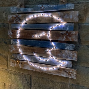 May include: A wooden wall hanging featuring a crescent moon shape formed by white lights. The moon is surrounded by small stars painted on the wood. The wood is painted in shades of blue and brown.