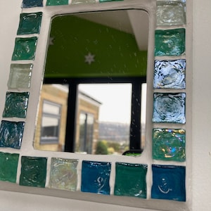 Op de afbeelding: Een witte spiegel met een moza&iuml;ekrand van blauwe, groene en iriserende glazen tegels. De spiegel reflecteert een uitzicht op een kamer met een groene muur en een raam.
