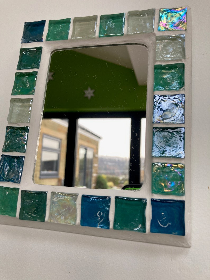 Op de afbeelding: Een witte spiegel met een moza&iuml;ekrand van blauwe, groene en iriserende glazen tegels. De spiegel reflecteert een uitzicht op een kamer met een groene muur en een raam.