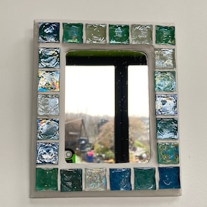 Op de afbeelding: Een kleine rechthoekige spiegel met een wit frame versierd met blauwe, groene en iriserende glazen tegels.