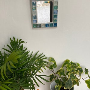 Op de afbeelding: Een kleine rechthoekige spiegel met een moza&iuml;eklijst gemaakt van blauwe, groene en witte tegels. De spiegel hangt aan een witte muur.