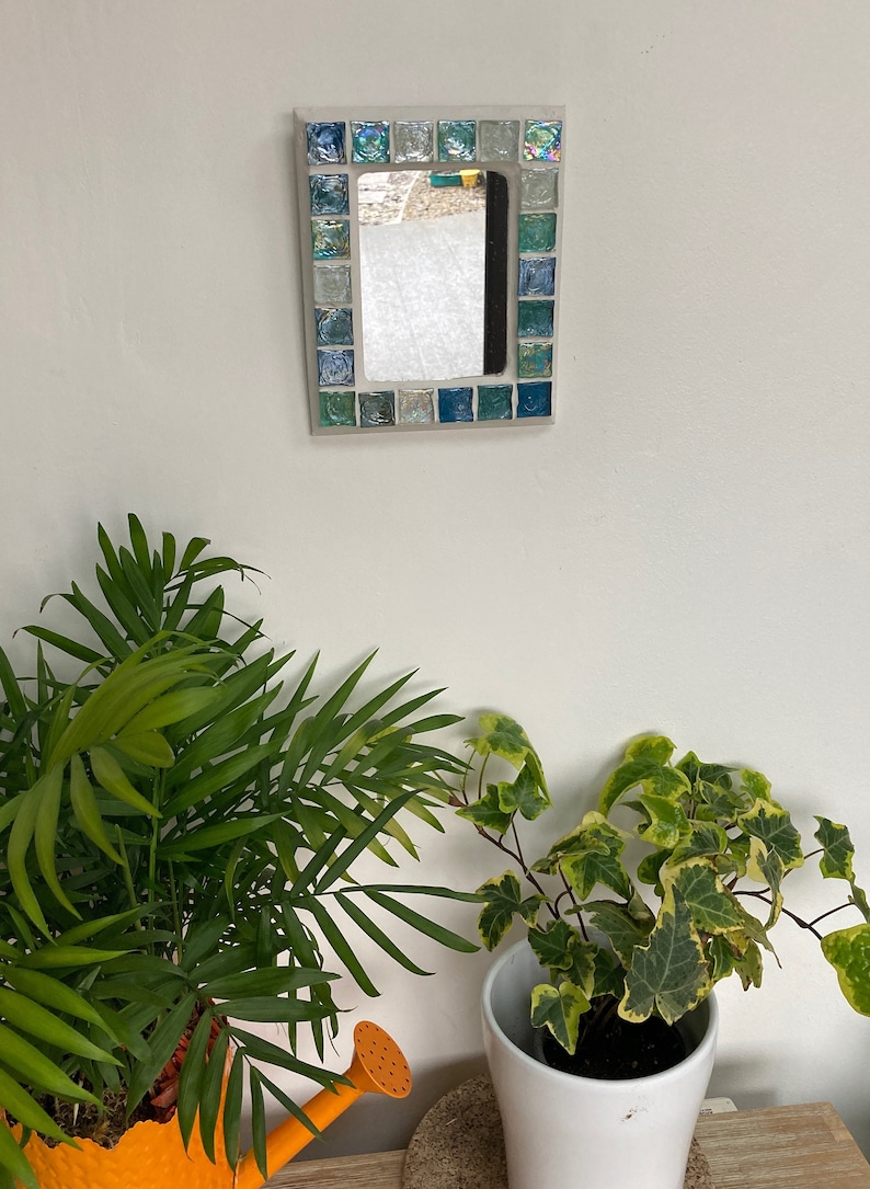 Op de afbeelding: Een kleine rechthoekige spiegel met een moza&iuml;eklijst gemaakt van blauwe, groene en witte tegels. De spiegel hangt aan een witte muur.