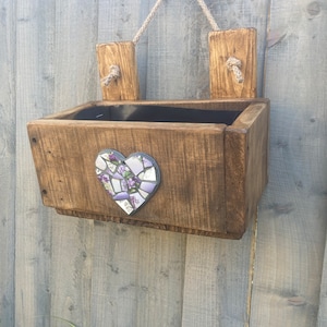 May include: A rustic wooden planter box with a heart-shaped mosaic design in shades of purple and white. The planter is hanging from a rope and has two wooden brackets on the sides.
