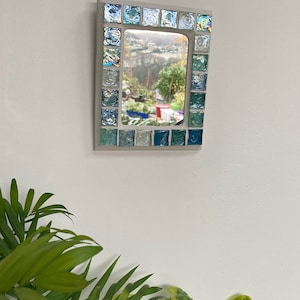 Op de afbeelding: Een kleine rechthoekige spiegel met een lijst van blauwe en groene moza&iuml;ektegels. De spiegel reflecteert een uitzicht op een tuin met bomen en een rood gebouw in de verte.