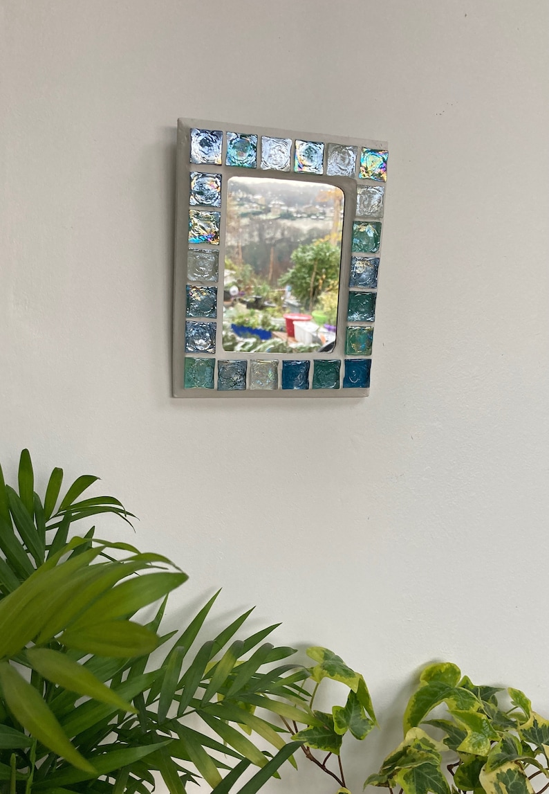 Op de afbeelding: Een kleine rechthoekige spiegel met een lijst van blauwe en groene moza&iuml;ektegels. De spiegel reflecteert een uitzicht op een tuin met bomen en een rood gebouw in de verte.