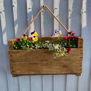 May include: A rustic wooden planter box with a rope hanger, filled with colourful flowers and green ivy. The planter is hanging on a white wooden wall.