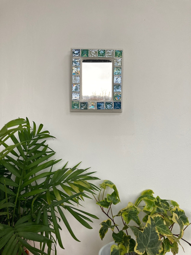 Op de afbeelding: Een kleine vierkante spiegel met een lijst van blauwe en groene moza&iuml;ektegels. De spiegel hangt aan een witte muur.