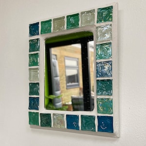 Op de afbeelding: Een kleine, vierkante spiegel met een wit frame versierd met groene en blauwe moza&iuml;ektegels.