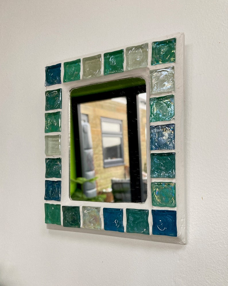 Op de afbeelding: Een kleine, vierkante spiegel met een wit frame versierd met groene en blauwe moza&iuml;ektegels.