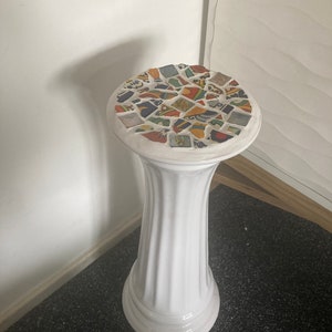 May include: A white ceramic pedestal with a mosaic tile top. The pedestal has a fluted column design and a round base. The mosaic top features colourful, irregularly shaped tiles in various colours.
