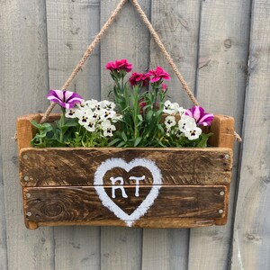 May include: A rustic wooden planter box with a heart painted on the front, featuring the initials "RT". The planter is hanging from a rope and filled with pink and white flowers.