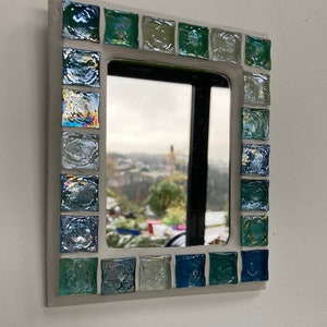 Op de afbeelding: Een kleine, wit geframed spiegel met een moza&iuml;ekrand van iriserende blauwe en groene tegels. De spiegel reflecteert een uitzicht op een stad en bomen.