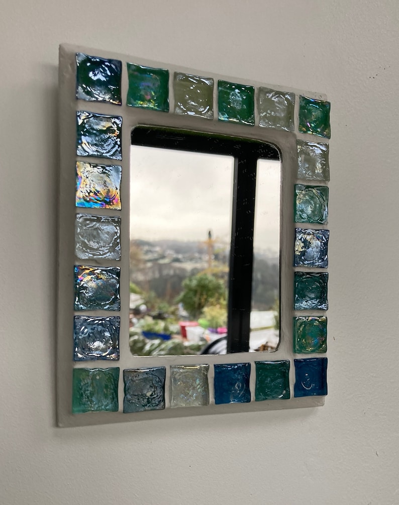 Op de afbeelding: Een kleine, wit geframed spiegel met een moza&iuml;ekrand van iriserende blauwe en groene tegels. De spiegel reflecteert een uitzicht op een stad en bomen.