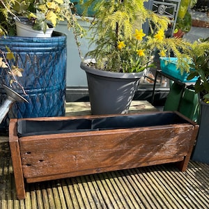 May include: A long, rectangular wooden planter box with a dark brown stain. The planter has two compartments and is sitting on a wooden deck.