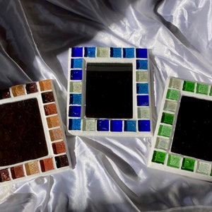 Op de afbeelding: Drie vierkante, met moza&iuml;ek ingelijste spiegels met verschillende gekleurde glazen tegels. De spiegels liggen op een witte stoffen achtergrond. De lijsten zijn bruin, blauw en groen.