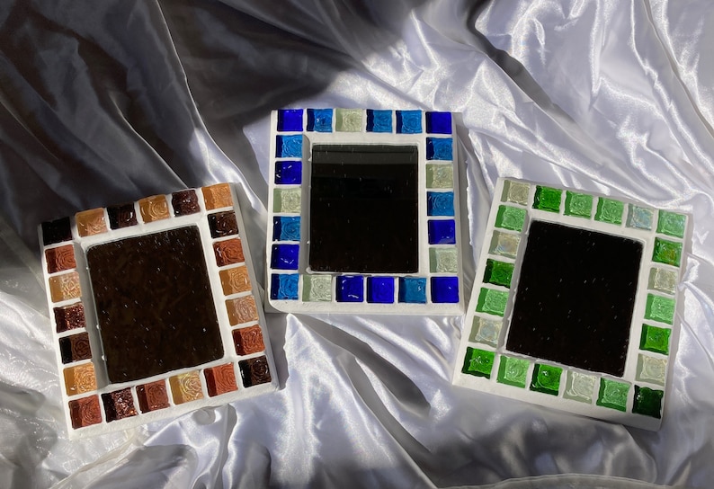 Op de afbeelding: Drie vierkante, met moza&iuml;ek ingelijste spiegels met verschillende gekleurde glazen tegels. De spiegels liggen op een witte stoffen achtergrond. De lijsten zijn bruin, blauw en groen.