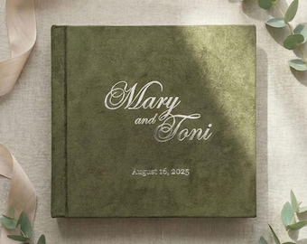 May include: A square, olive green photo album with the names "Mary and Toni" embroidered in white script. The date "August 16, 2025" is also embroidered in white. The album is on a neutral linen surface with eucalyptus and a cream ribbon.