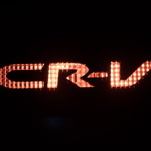 以下が含まれることがあります： グリッド状のライトで照らされた赤い「CR-V」ロゴ