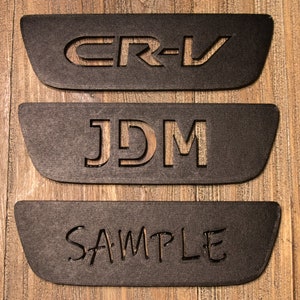 以下が含まれることがあります： 中央に「CR-V」、「JDM」、「SAMPLE」の文字が切り抜かれた、黒い長方形のプレートが3枚あります。