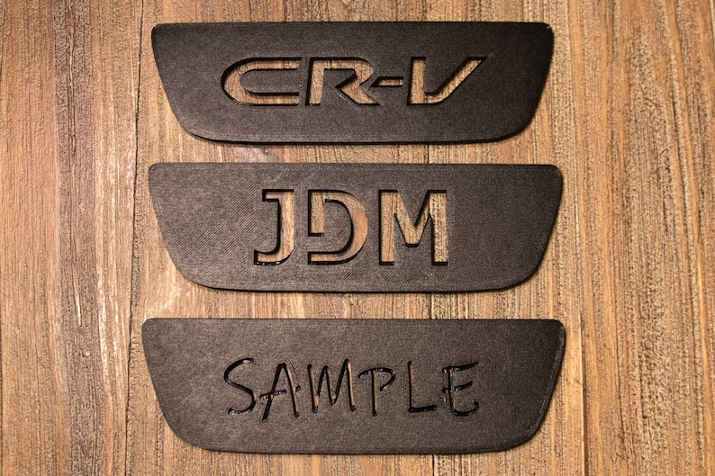 以下が含まれることがあります： 中央に「CR-V」、「JDM」、「SAMPLE」の文字が切り抜かれた、黒い長方形のプレートが3枚あります。