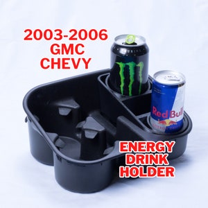 May include: Black plastic energy drink holder with four cup holders. The holder is designed to fit in a car's center console. Two energy drink cans are in the holder, one Monster Energy and one Red Bull.