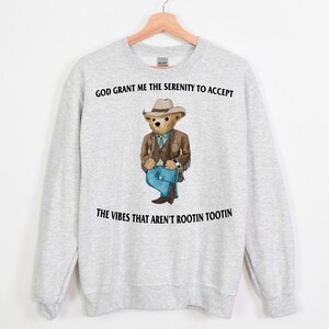 Könnte beinhalten: Ein graues Sweatshirt mit einem Aufdruck eines Cartoon-Bären, der einen Cowboyhut und Jeans trägt. Der Text "God grant me the serenity to accept the vibes that aren't rootin tootin" ist auf dem Sweatshirt aufgedruckt.