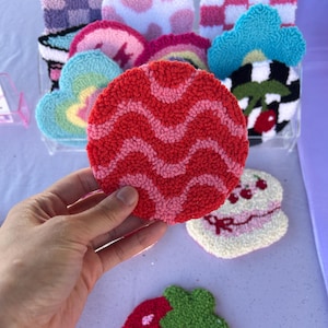 May include: A hand holds a red and pink wavy patterned coaster. Other colorful, tufted coasters in cloud, rainbow, and strawberry shapes are visible. The coasters are displayed in a clear container and on a light purple surface.