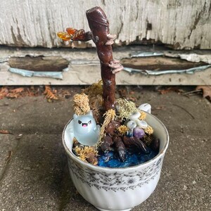 May include: A white porcelain teacup containing a miniature diorama with a brown twig, a translucent light blue ghost figurine with pink eyes, dried moss, small white mushrooms, and blue resin at the base, all arranged inside the cup.