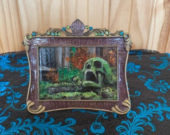 May include: A framed artwork featuring a green skull with an open mouth, set in a forest scene with orange and green foliage. The frame is ornate, with a brown and gold finish, and embellished with green and blue accents.