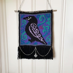 May include: A crocheted wall hanging featuring a black raven with purple and green details. The hanging has a black fringe and a silver charm.