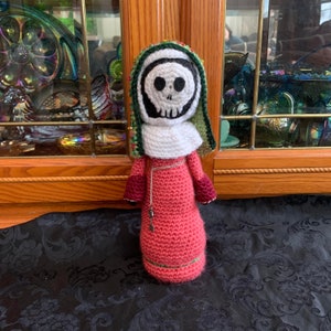 Puede incluir: Una muñeca de crochet con cara de calavera, vestida con una túnica con capucha verde y blanca y un vestido rosa. La muñeca sostiene una cadena de plata con un dije de llave.
