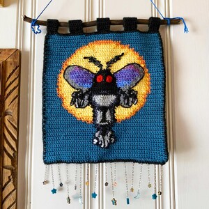 May include: A crocheted wall hanging featuring a black moth with red eyes and purple wings against a yellow moon on a blue background. The hanging has a fringe of silver chains with star and moon charms.