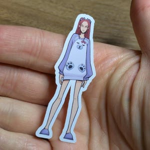 May include: A sticker featuring a cartoon character in a light purple bear-themed outfit. The character has red hair and is wearing a long-sleeved top with bear paw details and a bear face on the front. The sticker is held in a hand.