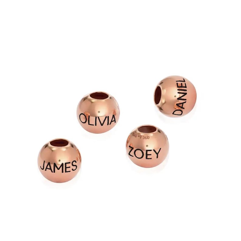 K&ouml;nnte beinhalten: Vier ros&eacute;goldene Perlen mit schwarzer Beschriftung. Die Perlen buchstabieren die Namen "James", "Olivia", "Zoe" und "Daniel".