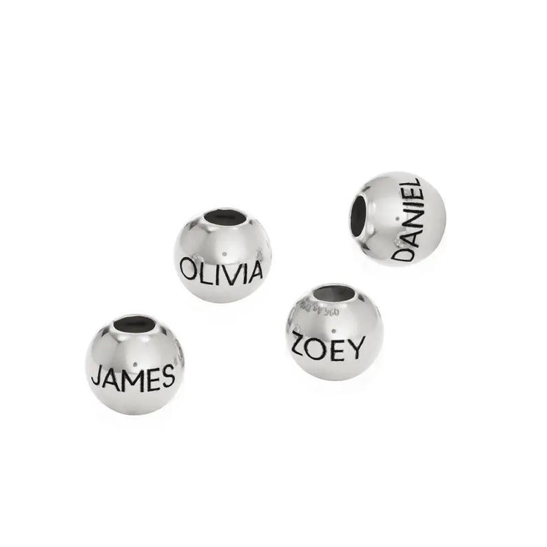 K&ouml;nnte beinhalten: Vier silberne Perlen mit schwarzer Beschriftung. Die Perlen sind mit den Namen "Olivia", "James", "Daniel" und "Zoey" graviert.