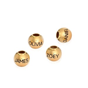 K&ouml;nnte beinhalten: Vier goldfarbene Perlen mit schwarzer Beschriftung. Die Perlen sind mit den Namen "Olivia", "James", "Daniel" und "Zoey" graviert.