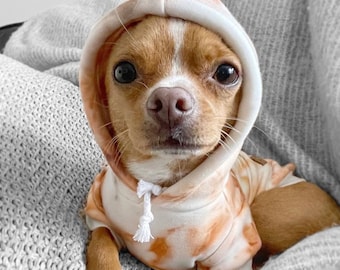 May include: A small brown and white chihuahua wearing a light brown and white tie-dye hoodie. The dog is sitting on a gray blanket.