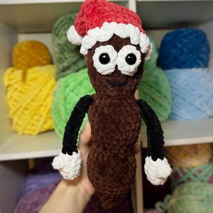 Puede incluir: Un juguete de peluche de color marrón tejido a crochet con forma de caca que lleva un gorro de Papá Noel tejido a crochet de color rojo y blanco. El juguete tiene brazos tejidos a crochet de color negro y manos tejidas a crochet de color blanco.